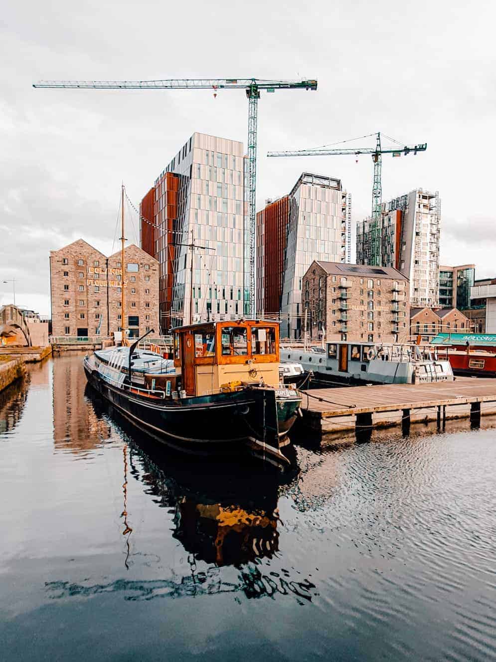 A scene at Dublin's Grand Canal Dock, showing modern high-rise buildings under construction, old industrial buildings, and boats moored along the water.