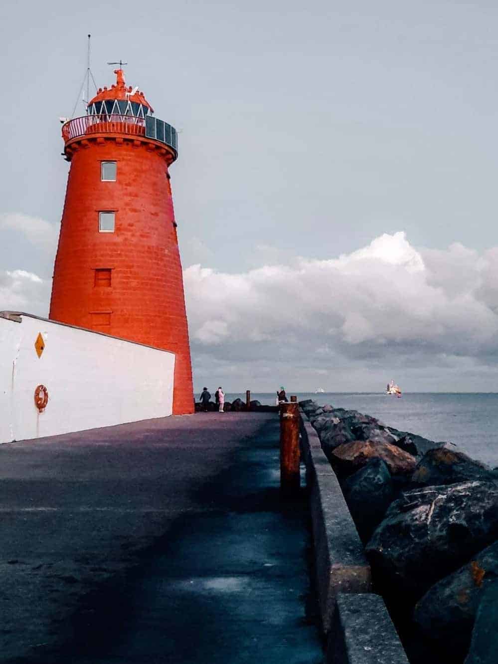 The iconic red Poolbeg Lighthouse in Dublin, standing at the end of a long pier, with people walking towards it and the sea on either side.