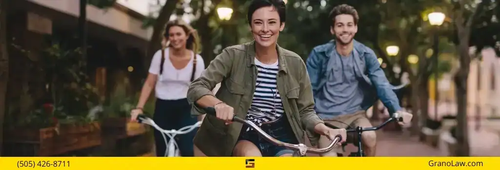 Three young adults smiling and riding bicycles down a tree-lined street, with the Grano Law Offices, P.C., logo and phone number overlay.