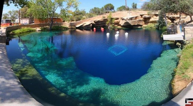 A wide-angle view of the Blue Hole in Santa Rosa, New Mexico, a deep, clear pool of water surrounded by rock formations and trees.