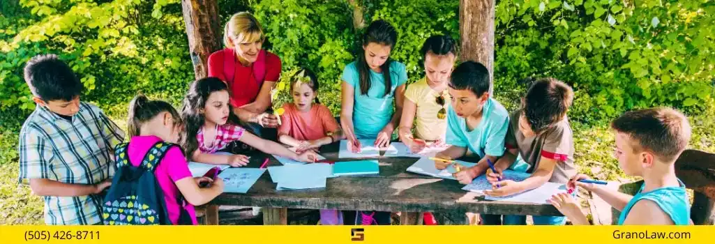 A group of children and a teacher drawing together at a picnic table outdoors in a sunny park, with the Grano Law Offices, P.C., logo and phone number.