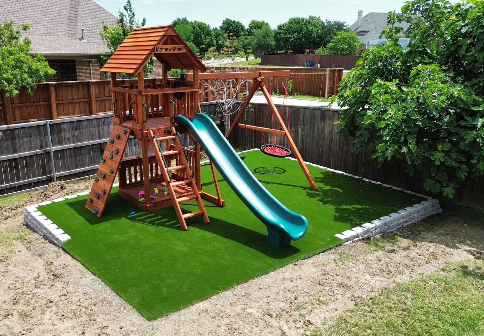 justin, texas, redwood swingset, Backyard playground with wooden Fort Davis playset featuring a green slide, rock climbing wall, swing set, and artificial turf base, surrounded by a wooden fence and landscaping.