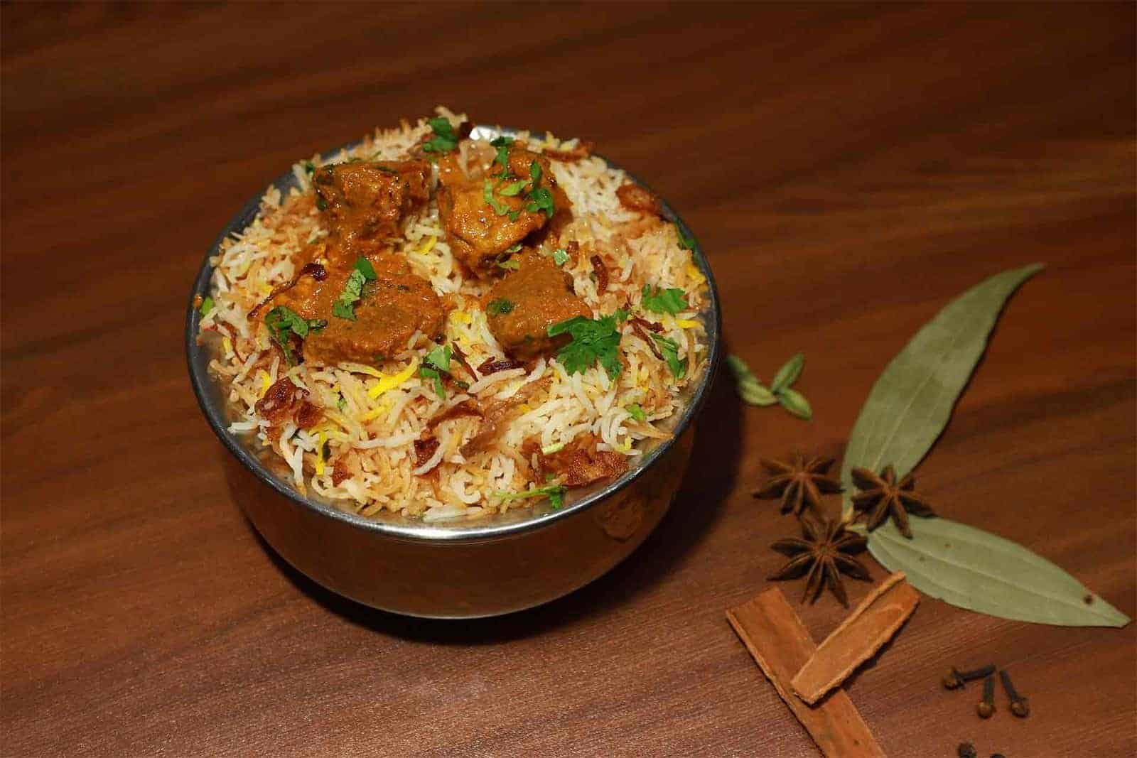 Fragrant Indian chicken biryani served in a steel bowl garnished with fresh cilantro, accompanied by whole spices on a wooden surface.