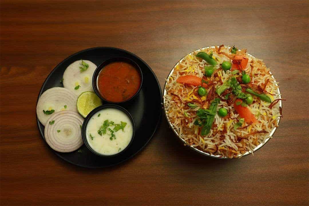 Steaming plate of flavorful biryani rice garnished with vegetables and herbs, served with side condiments and cooling yogurt sauce, highlighting authentic Indian cuisine.