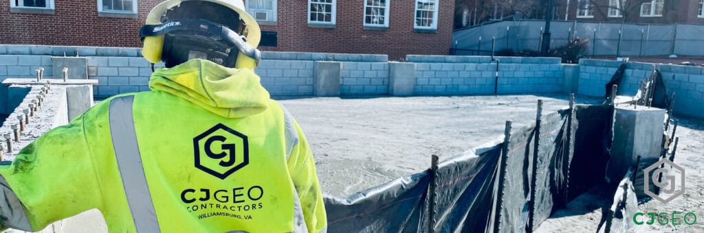 a worker wearing a high-vis jacket with CJGeo logo standing in front of silt fence containing a cellular concrete placement, with a brick academic building in the background.