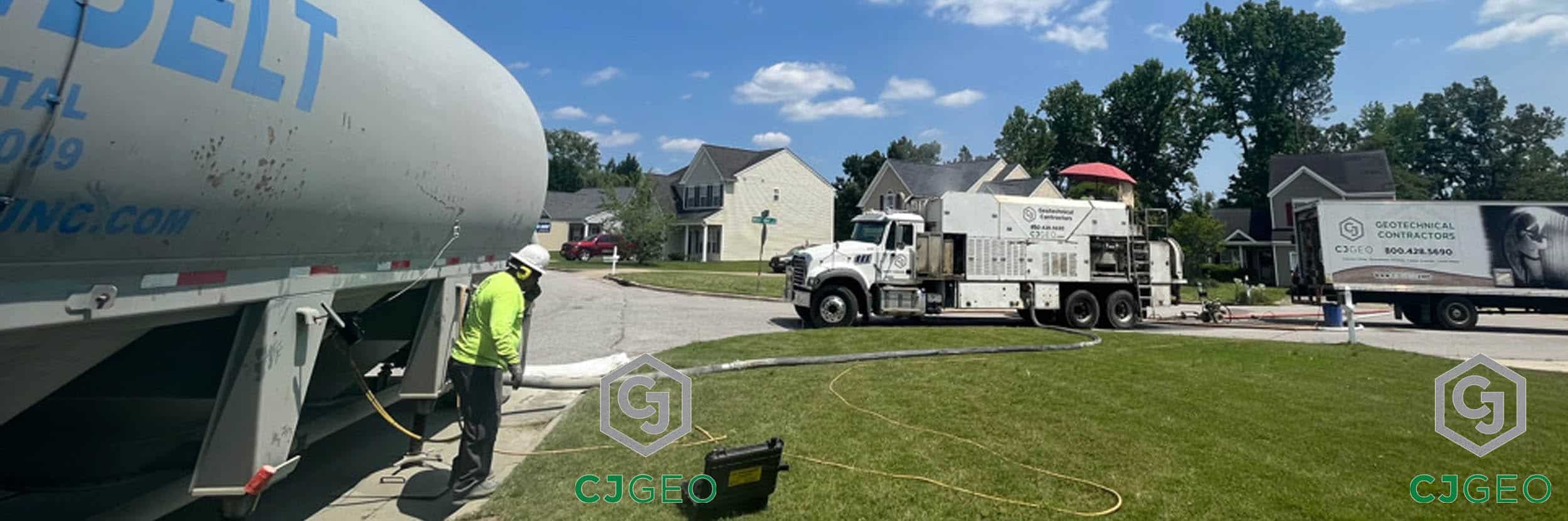 A CJGeo crew member operates a pump hose connected to a bulk cement truck, preparing to fill a grout mixer truck parked on a residential street.