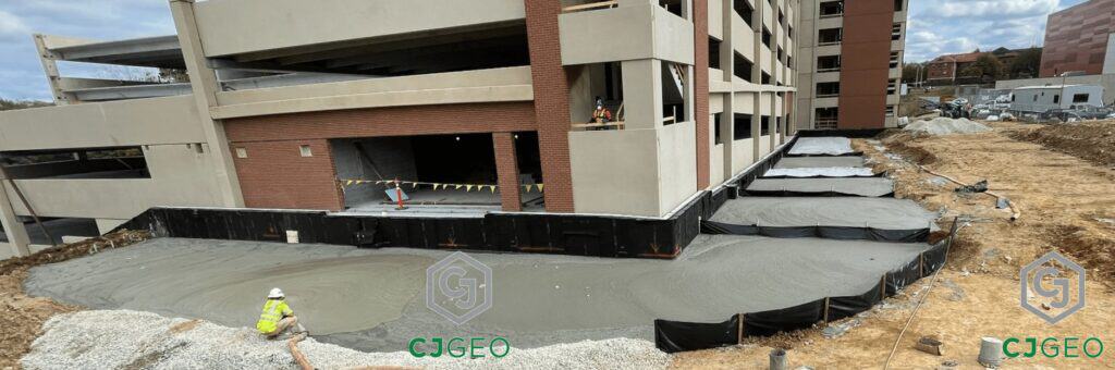 A CJGeo crew member uses a hose to place lightweight cellular concrete as structural backfill in a terraced excavation alongside a multi-story building foundation.