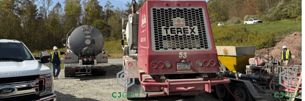 Grouting equipment and crew on a pipeline abandonment project. A large red TEREX concrete truck with number 53 on the back is visible, along with a grey tanker trailer, a white Ford pickup truck, and two crew members wearing hard hats and high-visibility vests. The work is taking place on a dirt and gravel site with trees in the background, demonstrating CJGeo's utility grouting services.