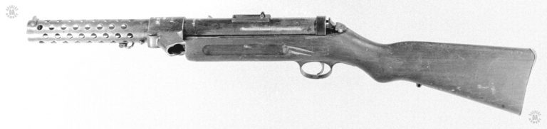 This is a left side view of the Bergmann MP 18/I, considered the first true submachinegun of World War I. The marking identifying the manufacturer are filled out in white on the receiver, underneath and past the back of the rear sight. The weapon is unloaded with no magazine in the distinctive rear-slanted magazine well just ahead of and above the stock. The unusual mouth of the magazine well is so that it can accept the 32-round “Snail-drum” of the Artillery Luger. At the back of the receiver is the spring-loaded plunger that would be pressed in to release the receiver from the stock. When released, the receiver would hinge upward so that the internal components could be removed and the barrel would be accessible for cleaning. PHOTO CREDIT: US Army