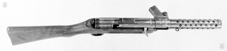 An upper right side view of the Bergmann MP18/I. On the top of the magazine housing can be seen the serial number and markings for the weapon. The serial number is repeated on the receiver tube just behind the magazine housing. The markings show the German style of identifying the weapon as the M.P. 18, I. The date (1920) underneath the name indicates that this weapon was issued to the German police after World War I. The hooked cocking lever on the right of the receiver is in the forward (uncocked or fired) position. At the rear of the slot the cocking lever travels in, is the safety notch. The lever would be pulled to the rear to cock the weapon for firing and lifted into the notch, where it would move slightly forward, and be locked into the “Safe” position. The serrated take down plunger Is visible at the rear of the receiver. Just below the magazine housing, in front of the stock, is the slotted head of the receiver hinge. PHOTO CREDIT: US Army
