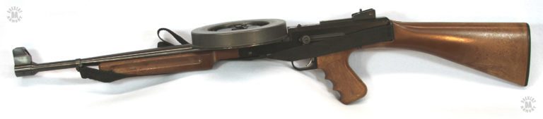 Left side view of the American 180. The cocking knob is visible just above the trigger and sling. Above the cocking knob, on the top of the receiver, is the magazine release. The very tall front sight assembly is to clear the sight line above the magazine. The central blade of the front sight, not visible in this picture, is protected by the two large wings on either side of it. PHOTO CREDIT: J. Purtillo