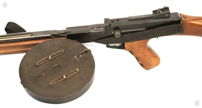 The unloaded American 180 with the bottom of the magazine next to it. The feed mouth of the empty magazine is clearly seen in this picture. On the barrel, just in front of the flat section. Is the forward magazine lock. The protrusion on the front of the magazine drum fits up under the notch to help secure the magazine in place. The center square indent on the bottom center of the magazine fits over the square lug just at the front of the receiver. PHOTO CREDIT: J. Purtillo