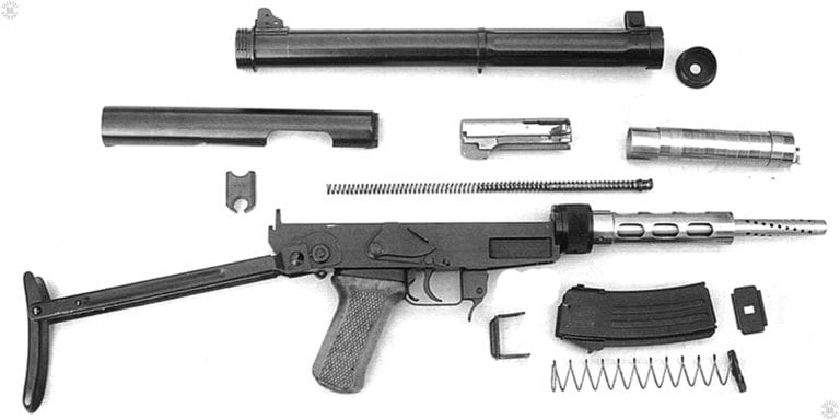 This is the Chinese Type 64 suppressed submachinegun stripped down to its basic components. At the top of the illustration is the long suppressor body that fits over the perforated and slotted barrel of the weapon. Above the barrel is the stack of baffles to the suppressor. Behind the baffle stack is the bolt above the mainspring. Below the top cover at the left in the picture, behind the mainspring, is a plastic buffer that minimizes the impact of the bolt against the back of the receiver. At the lower right is a 20-round magazine also stripped to its component parts for cleaning. PHOTO CREDIT: PRC