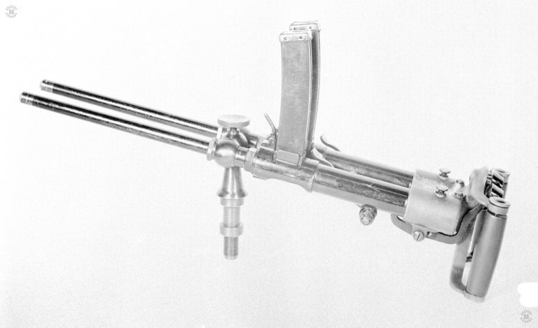 A left-side view of the Villar Perosa sitting on its central pedestal. The pedestal could be attached to a number of mounts including a small ground tripod, a carrying tray, and a bicycle. The twin vertical magazines are loaded into place and their individual magazine catches are the small bent rod in front of the magazine. Just behind and to the right of each magazine is the cocking lever for that individual firing mechanism. The large rectangular buttons at the rear of the weapon, between the vertical spade grips, are the trigger for each firing mechanism. The long lever underneath the weapon is the linkage from the left-hand trigger to the sear. PHOTO CREDIT: US Army
