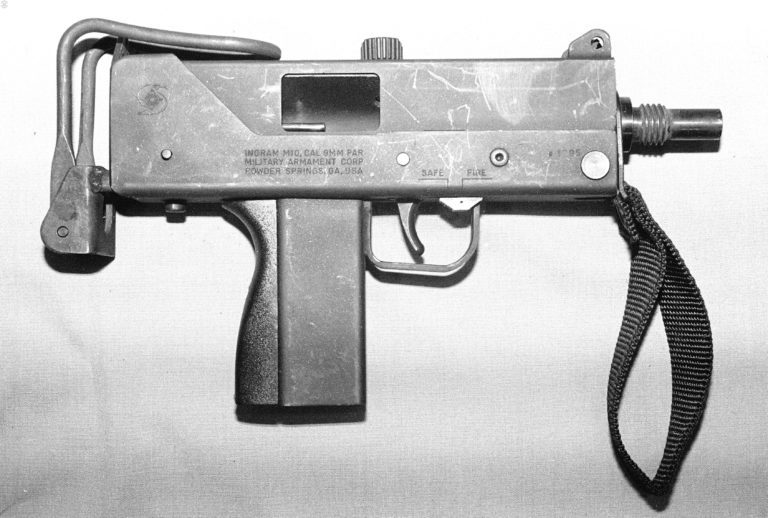 The right side of an Ingram M10 9mm with the bolt cocked but no magazine in place. The button extending from the bottom rear of the lower receiver is the stock latch. The rear portion of the stock has the bottom two legs squeezed together to release the butt and it is folded out and down. Then the stock catch is pressed in and the stock can be extended. The sliding safety switch can be seen just in front of the trigger on the outside of the trigger guard. The safety has been slid forward into the Fire position. With the bolt cocked to the rear, the tip of the ejector rod can be seen inside the ejection port. The nylon front strap is attached to the strap bar at the front of the weapon. PHOTO CREDIT: Kevin Dockery