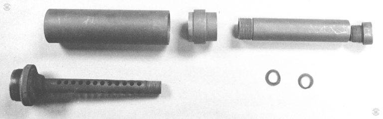 This is the M3 Greasegun suppressor assembly broken down partially for maintenance. The perforated barrel assembly is at the lower left, with the large tube of the first stage expansion chamber above it. The wire screen roll that would surround the barrel inside the expansion chamber tube is not in this picture. In front of the expansion chamber tube is the adapter plug that is threaded to go onto the muzzle of the barrel. The expansion tube is held in place by the adapter plug compressing it down onto the barrel bushing which is cut to accept it. The threaded tube to the right of the adapter bushing is the front tube that acts as a muffler device. Below the tube are two (used) screen washers from the over 200 that would fill the front tube assembly. Just to the right of the front tube is the threaded muzzle cap that seats down into the front tube and completes the assembly. This specimen is made of particularly thick and heavy steel components and may be a field manufactured expedient. PHOTO CREDIT: Kevin Dockery