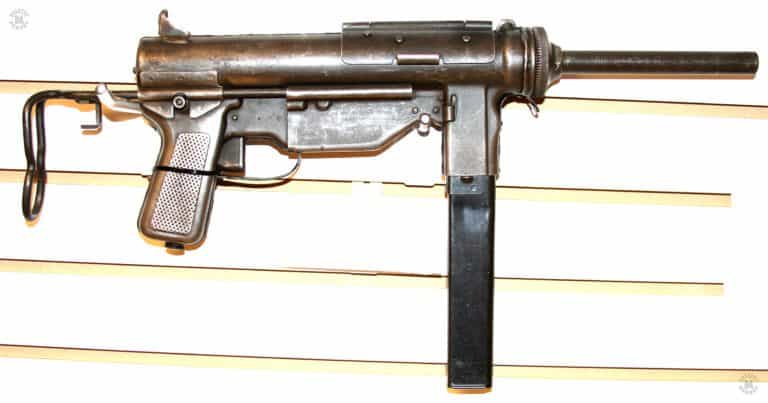 The left side of an M3A1 submachinegun. The markings of the weapon are visible on the side of the magazine well, this weapon having been made by the Ithaca Gun Company. There is no oiler clip on the side of the housing assembly as that style of oiler was replaced by the oil bottle in the grip. The stock is slid forward in the fully collapsed position. The L-shaped loading tool on the stock is up against the rear of the receiver. Sling mounts are welded to the side of the receiver. PHOTO CREDIT: Kevin Dockery