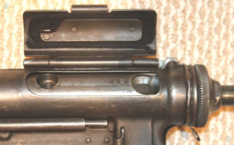 The open ejection port of the M3A1. The bolt is in the forward position and the ejection port cover is open. The sheet metal lug riveted to the inside of the ejection port cover is the safety. With the bolt in this position, closing the cover will engage the lug with the rear hole on the bolt. That locks the bolt in position against movement while handling. The large front hole in the bolt is for the finger of the operator to cock the weapon. PHOTO CREDIT: Kevin Dockery