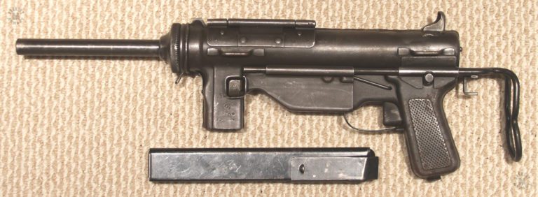 Left side of an M3A1 submachinegun with the stock collapsed. The 30 round magazine is laying under the weapon. The heavy reinforcement on the upper part of the magazine is necessary to strengthen the feed lips from the pressure of the round column. PHOTO CREDIT: Kevin Dockery