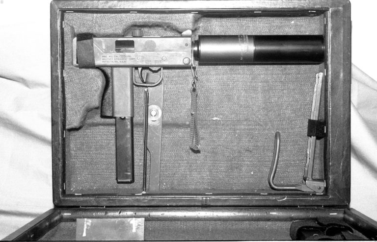 One of the most concealed manners of carrying an Ingram submachinegun, the Operational Briefcase. The fitted case will securely hold a loaded Ingram M11 submachinegun with the suppressor fitted and the sliding stock removed. In this specimen, the stock is secured to the side wall of the briefcase by a Velcro strap. The M11 is cocked and there is space in the fitted portion of the case to allow the bolt handle to move freely. The muzzle of the suppressor butts up against the side of the case where there is a hole through the wall of the case to allow for passage of the projectiles. On the outside of the case there is a small frame covering the exit hole for the projectiles. The frame allows for a standard business card to be slipped into it to cover the hole. The long spring-loaded bar extending from the trigger to the outside of the bottom of the case is the trigger for the Operational Briefcase. The L-shaped extension at the top of the bar bears against the trigger of the submachinegun. To fire, the case would be pulled up under the arm of the shooter and the trigger bar extending out of the bottom of the case be pushed forward. The spring will pull the trigger bar off of the submachineguns trigger when it is released. There were also specimens of the Operational Briefcase built to hold the M10 submachinegun, in either .45 ACP or 9x19mm. Those weapons had to be mounted with a special shorter suppressor in order to fit in the case. PHOTO CREDIT: Kevin Dockery