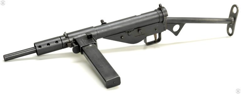 A left-side front view of the original configuration of the Sten Mk II. The locking mechanism for the magazine housing can be seen at the front side of the housing itself. The large half-disk is the plunger that is pulled out against spring pressure to unlock the housing and allow it to rotate downwards. The catch for the barrel nut is below the spring on the plunger. The L-shaped piece on the opposite side of the magazine well is the magazine catch itself. Pressing the catch down from the top releases the magazine from the well. PHOTO CREDIT: US ARMY