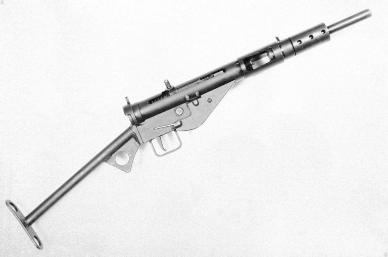 The right side of an early production Sten Mk II. The weapon is fitted with the No 2 Mk II “T” stock that was originally developed for this model Sten, but could later be found on any of the Marks. The selector switch is the round button just in front of the trigger guard on the flat side of the trigger mechanism. This specimen has had the button pressed in, setting the weapon for automatic fire as indicated by the large “A” on the end of the switch. The bolt has been drawn fully to the rear and lifted up into the safety notch. This specimen has the Mk II cocking handle, recognizable by its rounded end, knurled sides, and large integral “washer” at the base of the exposed portion. With the bolt drawn back, the entire ejection port and magazine well is open in this picture. The small protrusion on the back side of the ejection port is the fixed ejector. The front sight is the protrusion on top of the receiver in front of the magazine housing. PHOTO CREDIT: US ARMY