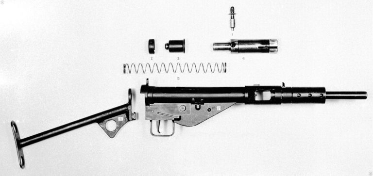 The component parts of a field stripped Mk II Sten, with the barrel assembly still attached to the receiver. Part #1 is the Mk II cocking handle. Part #2 is the Return spring cap that secures to the back end of the receiver. Part #3 is the return spring housing that sit over the end of the return spring and extends through the center of the return spring cap. It is this protrusion of the housing through the cap that helps secure the attachment of the stock to the weapon. Part #4 is the bolt, or Mk I Breech block as it is called officially. Part #5 is the Mk I Return spring that drives the bolt. Part #6 is the No 2 Mk II “T” stock. The lug on the front bottom of the stock seats inside the rear of the trigger mechanism and up into the extension on the rear of the receiver body. Part #7 is the Mk I Trigger mechanism cover, identifiable by the single screw that secures it to the bottom of the receiver. The receiver could be broken down further by the removal of the barrel assembly. Access to the trigger mechanism could be made by the removal of the trigger mechanism cover. PHOTO CREDIT: US ARMY