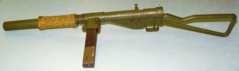 The left side of a Sten Mk II(S) loaded with a standard 32-round magazine. The weapon is fitted with the No 3 Mk I skeleton stock, which was formed from U-channel tubing. The bolt is drawn back and raised into the safety slot at the end of the cocking handle slot. The lacing for the securing of the canvas thermal sleeve is also visible on this side. PHOTO CREDIT: KEVIN DOCKERY