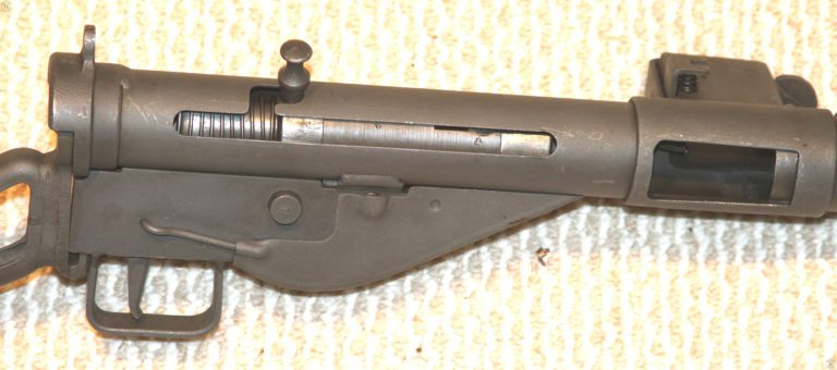 A closeup right side view of the Sten Mk II receiver. This weapon has been fitted with the Mk V cocking handle. The Mk V cocking handle was fitted to all Stens by orders issued on 15 April 1944. The long length of the Mk V Cocking handle is due to it being able to be pressed in, where it is secured by spring pressure, though the body of the receiver and bolt. A small stud on the bottom of the Mark V Cocking handle protrudes out from the bottom of the part and through a hole drilled for the purpose on the lower left side of the weapon’s receiver. The Mark V Bolt handle can lock the bolt in the forward, uncocked, position for safety.
     On this specimen, the bolt has been drawn back and the bolt handle lifted up into the safety slot. The weapon has been set on single shot or semiautomatic fire by pressing in the selector button from the left side of the weapon. The selector button, in the center of the trigger housing, has a stamped letter “A” in it to indicated that pressing in the button from this side would set the weapon on automatic fire. PHOTO CREDIT: KEVIN DOCKERY