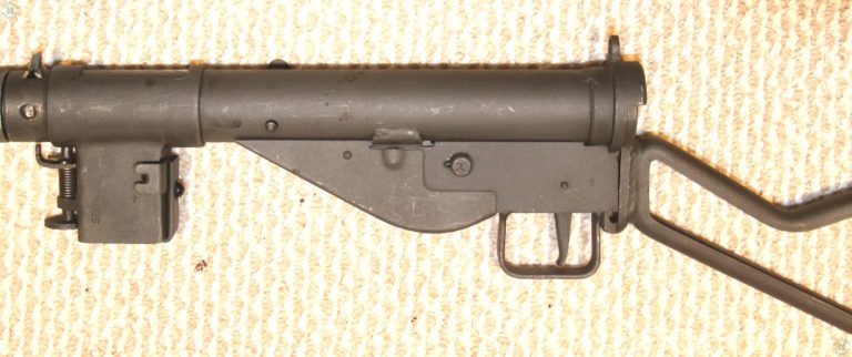 A left side view of a Mk II Sten submachinegun. This specimen has been fitted with the No 3 Mk I skeleton stock which is formed from U-channel steel. The selector switch has been pressed in, setting the weapon on “Repetition” or semiautomatic fire. Above the front of the trigger housing is the protruding end of the Mk 5 safety cocking handle. With the handle end protruding through the hole drilled for it, the bolt is locked in the forward position. The magazine housing has been rotated to the down position.
