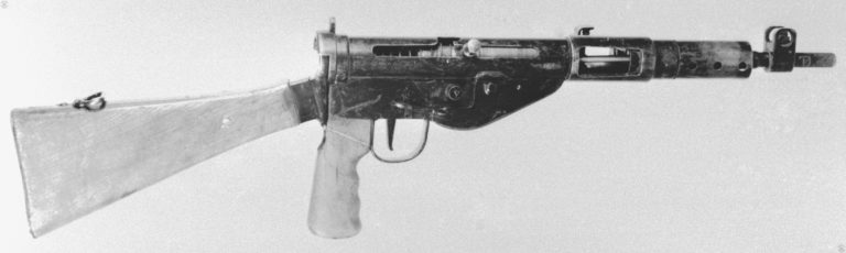 The right side of a Sten Mk V with the bolt in the cocked position. The trigger mechanism has been moved forward to allow the weapon to mount a rear pistol grip. The pistol grip has to have a section of the wood removed at the upper rear for it to clear the attachment of the wooden stock. This specimen is fitted with the alternative Mk3 buttstock with the slightly curved, almost flat, metal butt plate. The forward vertical grip is missing on this specimen. PHOTO CREDIT: US ARMY