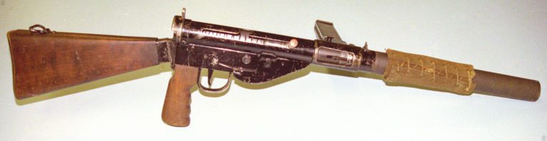 The right side of the Mk VI Sten. The front grip is not attached on the Mk VI due to the suppressor body. The front sight is moved back from the muzzle of the weapon to the top of the front of the receiver, in roughly the same position as on the Sten Mk II and Mk II(S). The body of the weapon has been given a coating of black paint while the suppressor is covered with a gray coating of phosphate. The canvas thermal cover overlaying a wrapping of asbestos string, is the same model as used on the Sten Mk II(S). The Mk 5 Safety cocking handle on the bolt is pressed in, locking the bolt in the forward position. PHOTO CREDIT: KEVIN DOCKERY