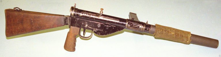 The right side of a Sten Mk VI with the bolt drawn back and lifted into the safety notch. The long length of the Mk 5 safety cocking handle can be seen as the part extends up from the bolt. This length is used when the bolt is in the forward (fired) position and the handle is pressed in to engage a hole on the opposite side of the receiver. The top of the empty magazine can be seen through the open ejection port. PHOTO CREDIT: KEVIN DOCKERY