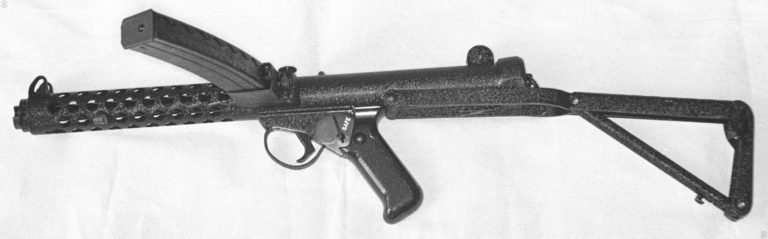 A left-side view of the Sterling L2A3 (Mk 4) submachinegun. The heavy crackle-paint finish and markings on the selector switch indicated that this is a Mark 4 version intended for international sale. The pistol grip is at the point of balance for the piece with the stock extended. At the top of the grip, behind the trigger, is the safety selector. In this position, the selector is easily operated with the shooter’s right-hand thumb. On this specimen, the selector is set on “1” indicating semiautomatic fire only. Pressed forward to the number “34”, the weapon is set to fire on full automatic. The stock has been fully unfolded and locked into position. The small hook-like projection on the bottom tube of the buttstock, just in front of the butt plate, is the butt plate catch that is pushed forward to unlock the tube and start the folding sequence. Near the muzzle of the weapon is the stud that acts as the standard for mounting a bayonet. The weapon is loaded with a 34-round magazine showing its distinctive forward curve to aid in feeding. PHOTO CREDIT: KEVIN DOCKERY