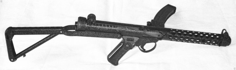 A right-side view of a Sterling Mk 4 submachinegun showing the crackle paint finish that is often found on the guns intended for commercial sale. The bolt is in the forward (uncocked) position. The helical cuts along the sides of the bolt can be seen through the cocking lever slot and the ejection port. These cuts aid in keeping the bolt clear of foreign matter that could cause a jam. Just visible near the muzzle of the weapon can be seen a second finger guard, this one intended to keep the firer’s hand from slipping forward while firing. PHOTO CREDIT: KEVIN DOCKERY