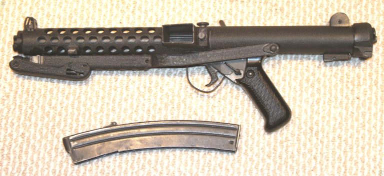 The left side of a Sterling L2A3 with the stock folded and the magazine removed. The selector switch behind the trigger is set to A for full automatic fire. The round button behind the magazine well is the outer part of the ejector. The large flat-topped knob on top of the weapon, just above and behind the ejection port, is the magazine release. The magazine is the standard Sterling 34-round model. PHOTO CREDIT: KEVIN DOCKERY