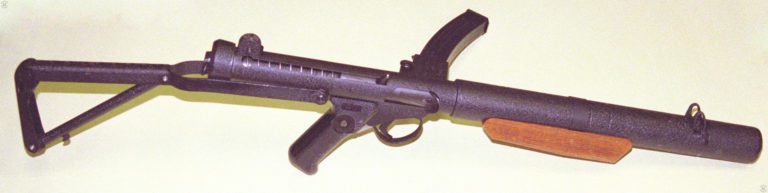 The upper right side of a Sterling Mk 5 submachinegun. The stock is filly extended and locked into place. The bolt is in the forward position and the wooden front grip is underneath the barrel. This specimen shows that the crackle finish used primarily on the Sterling commercial weapons does not show a reflective surface under normal lighting. PHOTO CREDIT: KEVIN DOCKERY