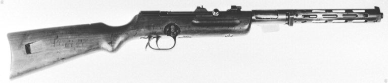 Right side view of an Erma EMP modified for late production. In this specimen a leftover submachine gun stock was modified to fit the Erma design. Missing is the very distinctive vertical foregrip of the standard Erma, in its place are deep finger grooves on either side of the forestock. The cutaway section of the stock, above the front of the trigger guard, is the selector switch. The bolt handle is in the forward (fired) position. On the top of the receiver, in front of the bolt handle, is the special bolt lock system that was placed on weapons issued to German police. The system would lock the bolt solidly in the forward position, preventing any accidental discharge if the weapon was dropped or struck solidly on the butt. The long lever behind the trigger guard is the “Verschluβhebel,” the locking lever stop which unlocks the receiver from the stock for field stripping. PHOTO CREDIT: US Army
