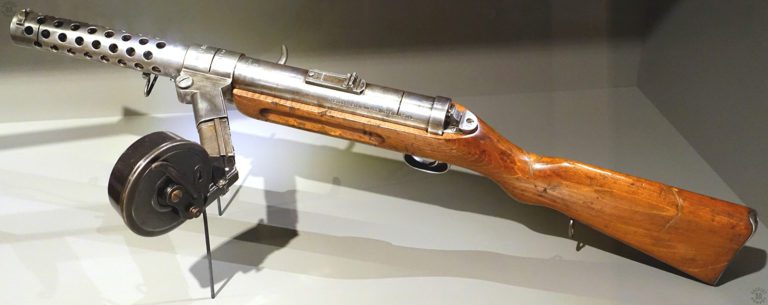 An upper left-side view of an MP18/I submachine gun, loaded with a late-model 32-round Luger drum magazine. The drum magazine is identified by the flat folding lever used to load the device. The first model drum had a telescoping round rod. The position of the curved bolt handle on the right side of the weapon indicates that the bolt is in the forward, uncocked, position. The rear sight on the top center of the round receiver has two folding leaves, one for 100 and one for 200 meters. The 200 meter leaf is in the folded position with the 100 meter leaf up for aiming. The serrated lug at the rear of the receiver would be pushed forward to unlock the receiver, allowing it to hinge open for cleaning. The three slit cuts across the muzzle, magazine well, and receiver, are to deactivate the weapon for display. PHOTO CREDIT: Braunschweigisches Landesmuseum