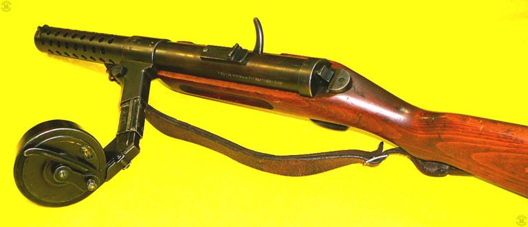 A left-rear view of an MP18 submachinegun fitted with a 32-round Late model Luger drum magazine. The spacer sleeve has been placed over the feed neck of the magazine, between the base section of the magazine and the magazine well. The bolt is in the rearmost, cocked, position as seen by the curved bolt handle on the right side of the receiver. The bolt handle has been lifted up into the safety notch in the upper side of the handle slot. On the center top of the receiver is the base for the rear sight with the 100-meter leaf up for aiming. At the rear of the receiver is the serrated surface of the receiver catch. The catch would be pressed in against spring pressure to unhook from the flat side of the retaining plate attached to the stock. With the receiver unlatched, the upper receiver would hinge up so that the internal parts can be removed for cleaning. PHOTO CREDIT: WARREN WHEATFIELD