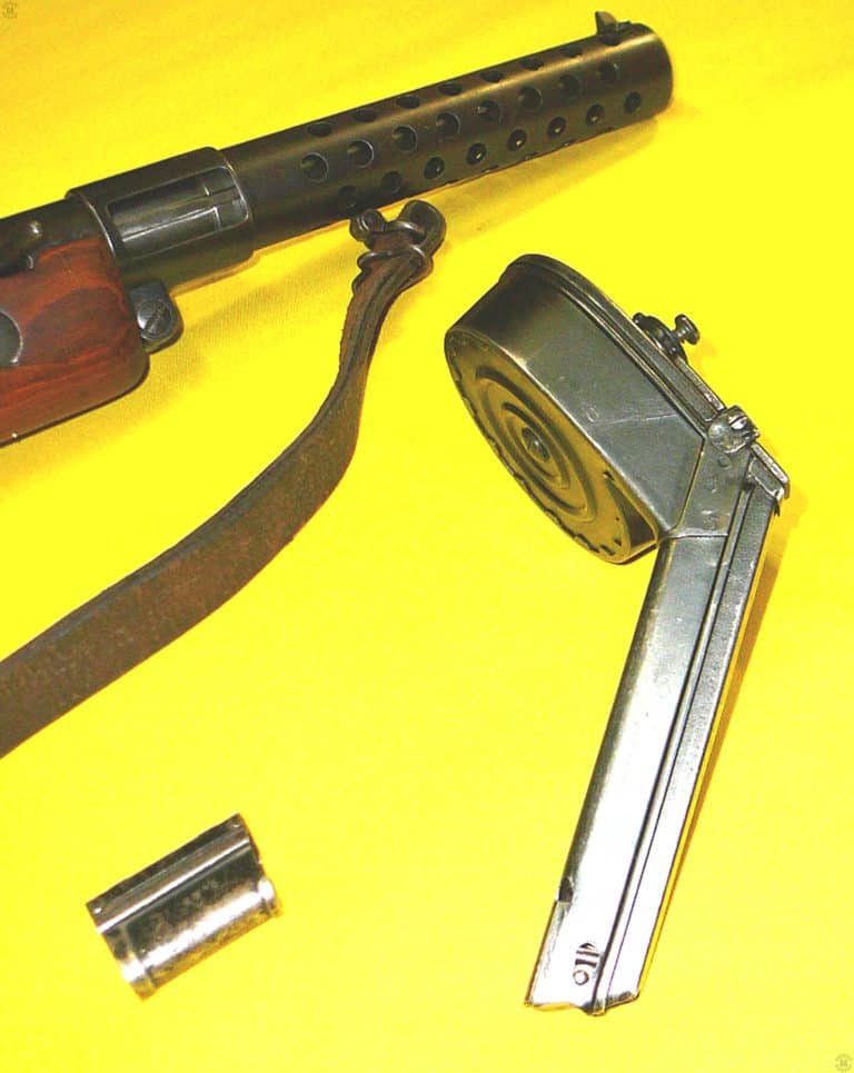The right side of the Late model luger drum magazine is uppermost in this picture. Loaded rounds would extend the length of the feed neck of the drum and continue around the circumference of the drum body. A very strong springs drives the column of rounds, requiring a special loading tool to properly fill the magazine. On the lower left is the spacer sleeve that makes using the drum magazine to load the MP18/I much easier and faster. Without the sleeve on the feed neck, the magazine could be carefully loaded into the weapon, the magazine catch seating in the small notch on the front edge of the feed neck, just below the top of the magazine. But in the rush of combat, the drum could easily be over-inserted and a jam result, taking the weapon out of action for a few moments. PHOTO CREDIT: WARREN WHEATFIELD