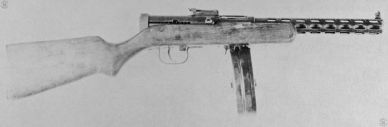 The specimen is of an earlier production model of the Model 1934/38. The cooling slots for air circulation on the barrel jacket make up 7 rows of 8 slots each, the slots being 7.9 mm (.031 in) wide and 23.6 mm (0.93 in) long. They are cut into the barrel jacket that is itself machined from solid stock. The bolt is in the forward position and locked in place. The safety on the PPD 34/38 is the sliding locking bar on the upper side of the cocking handle. It can be slipped into the square notch at the front or rear of the cocking handle slot, and lock the bolt in position. The rear (cocked) cocking handle slot can be seen on the upper side of the shot, above the trigger area. The flag-like lever in the front of the trigger is the fire selector lever, located on the right side of the weapon. The lever pivots on a pin at the front of the selector, as seen in this view. With the selector rotated to the rear, the weapon is set to fully automatic fire and the number 71 can be read on the right side of the flag. The number 73 was also used on some specimens. This weapon is loaded with the standard 71-round drum made for it. PHOTO CREDIT: US ARMY
