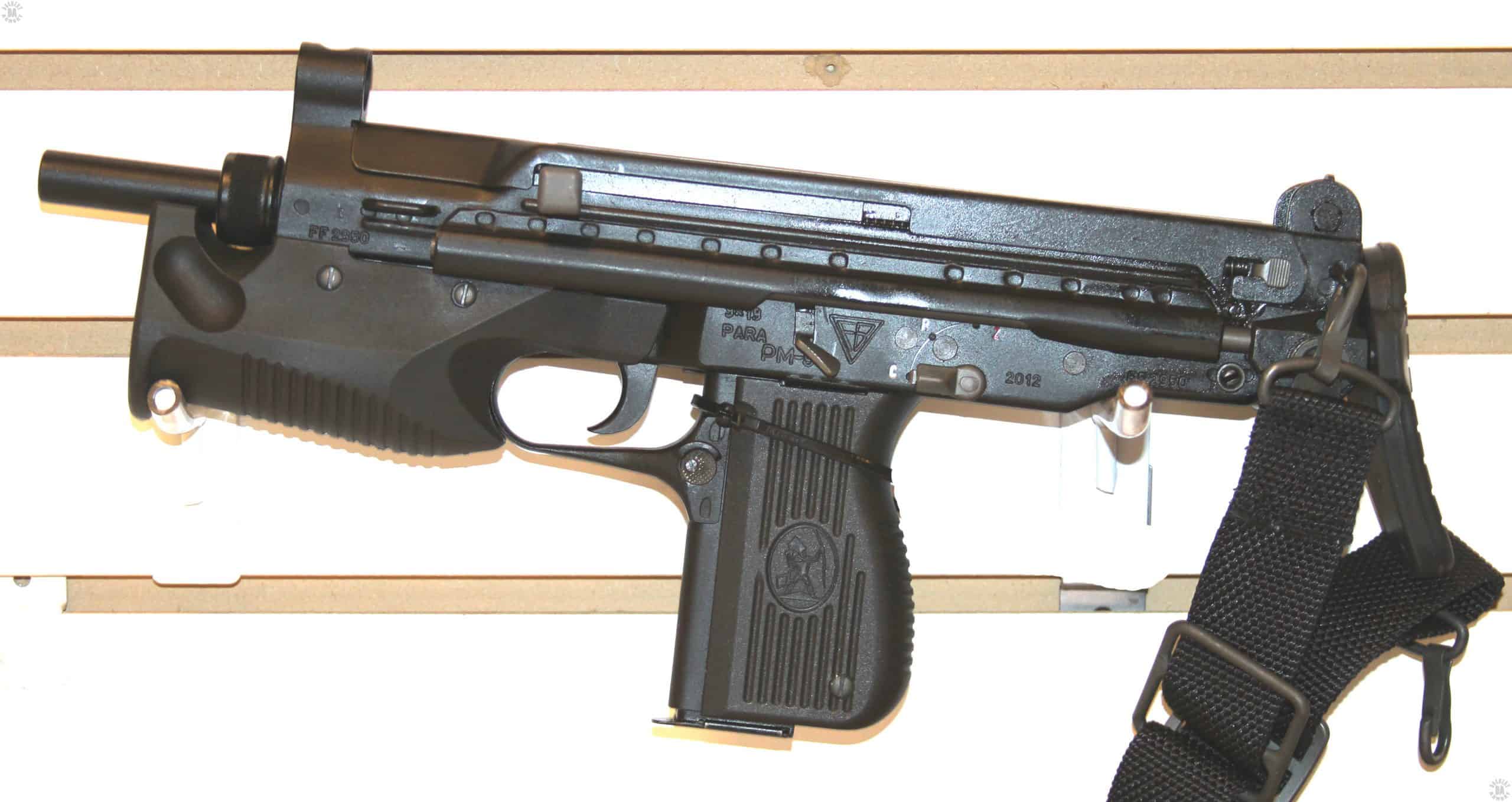 Left-side view of a PM-98. This specimen is fitted with a small cocking knob, the grey square at the front of the cocking knob slot, rather than the larger version that extends up above the top cover of the weapon. This is the cocking knob normally seen on the PM-06. The stock is fully forward in the collapsed position. The selector switch above and behind the pistol grip is set to “C” for Całkowicie automatyczny or Fully Automatic fire. The other markings are “P” for Półautomatyczny or Semiautomatic Fire. The top marking in red is a “Z” for Zablokowany or Locked. When the selector switch is straight up in the Locked position, the weapon is on safe and the bolt cannot move. The selector has to be in the P or C position in order to cock the bolt and chamber a round. This specimen is loaded with the 15-round magazine for maximum concealability. PHOTO CREDIT: KEVIN DOCKERY