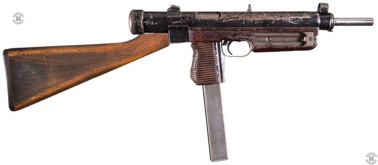 A right-side view of the fixed-stock 7.62x25mm Vz-24. This later-production specimen has the rear sight base and stock attachment point made of metal stampings welded to the receiver rather than the earlier production heavy machined parts. The receiver has been painted rather than blued during the production process, as can be plainly seen from the wear in the pain. The reddish furniture on the weapon is manufactured from a form of Bakelite plastic. The magazine loading tool is at the front side of the fore grip. Behind the trigger is the safety switch in the rearward, off, position. The bolt is in the forward position. This specimen is loaded with the 40-round magazine from the 9mm version of the series. The long magazine shows the angle the pistol grip has been changed to from the 9mm weapons. PHOTO CREDIT: ROCK ISLAND AUCTION
