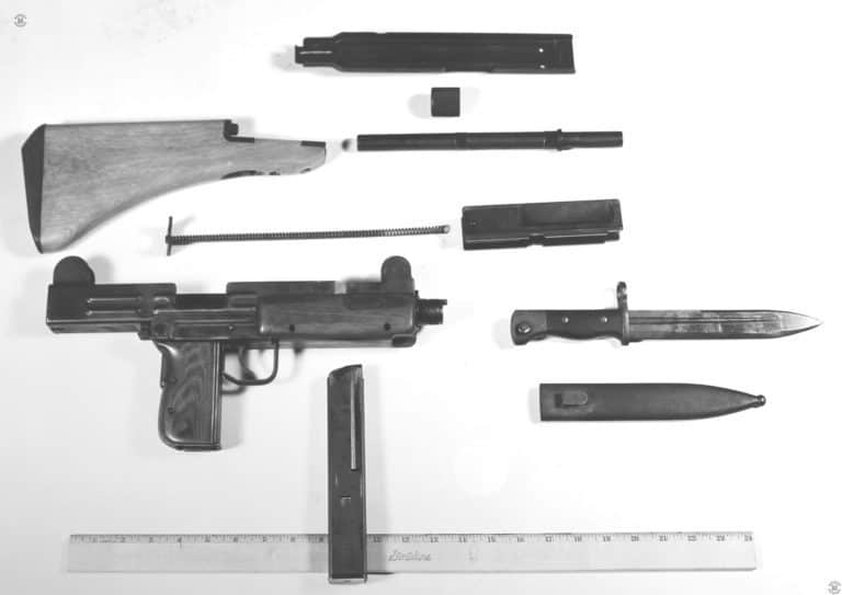 The right-side of a field stripped pre-production Uzi. At the top is the receiver cover, upside down with the cocking knob covered by the body of the part in this picture. Below the top cover is the barrel nut, below that the barrel itself. The use of the telescoping bolt allows for the Uzi design to have a relatively long barrel while still keeping the overall length compact. To the left of the barrel is the complete wooden stock assembly. Below the stock is the mainspring assembly. To the right of the mainspring is the bolt, the large hollow portion of which can be seen in the ejector opening. To the left is the receiver of the weapon, stripped as it would be for maintenance and cleaning. To the right of the receiver is the Uzi bayonet above its metal scabbard. At the bottom near center of the picture is the 30-round magazine. The magazine has been reversed in this picture and is facing to the rear of the weapon. PHOTO CREDIT: US NAVY HISTORICAL ARCHIVES