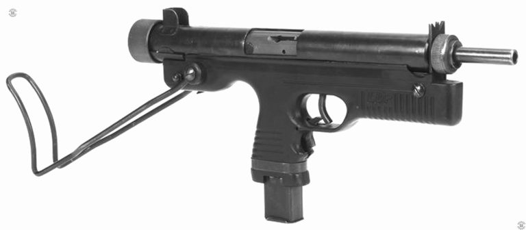 A right-front view of the Kommando carbine with the stock in the open (extended) position. The grip safety is visible underneath and behind the trigger guard. The spring-loaded grip safety is pulled back to allow the trigger to be pulled and the weapon fired.
