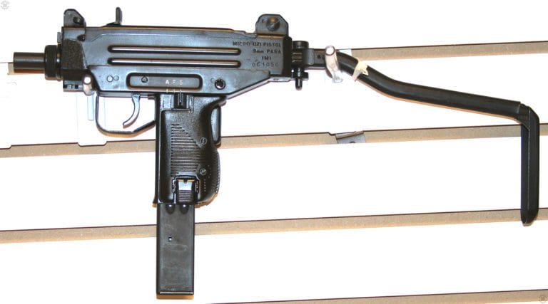 The left side of a Micro-Uzi, converted from the Uzi Pistol by IMI. The extended stock has a rubber coating rather than the more common early leather covering.  The front barrel nut is the shorter, more compact version used on the Micro-Uzi. On the front of the receiver, just ahead of the suspension hook of the display, is the bayonet lug used for the attachment of accessories such as a vertical grip. The short barrel on this specimen does not have the two angled cuts on the top and right side of the muzzle that act as a compensator. The safety/Selector is set to Safe in this illustration and the specimen is loaded with a 32-round magazine. PHOTO CREDIT: KEVIN DOCKERY