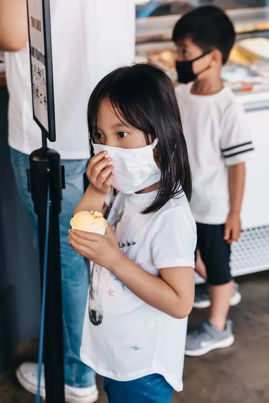 A child wearing a mask is holding an ice cream cone while standing in line.