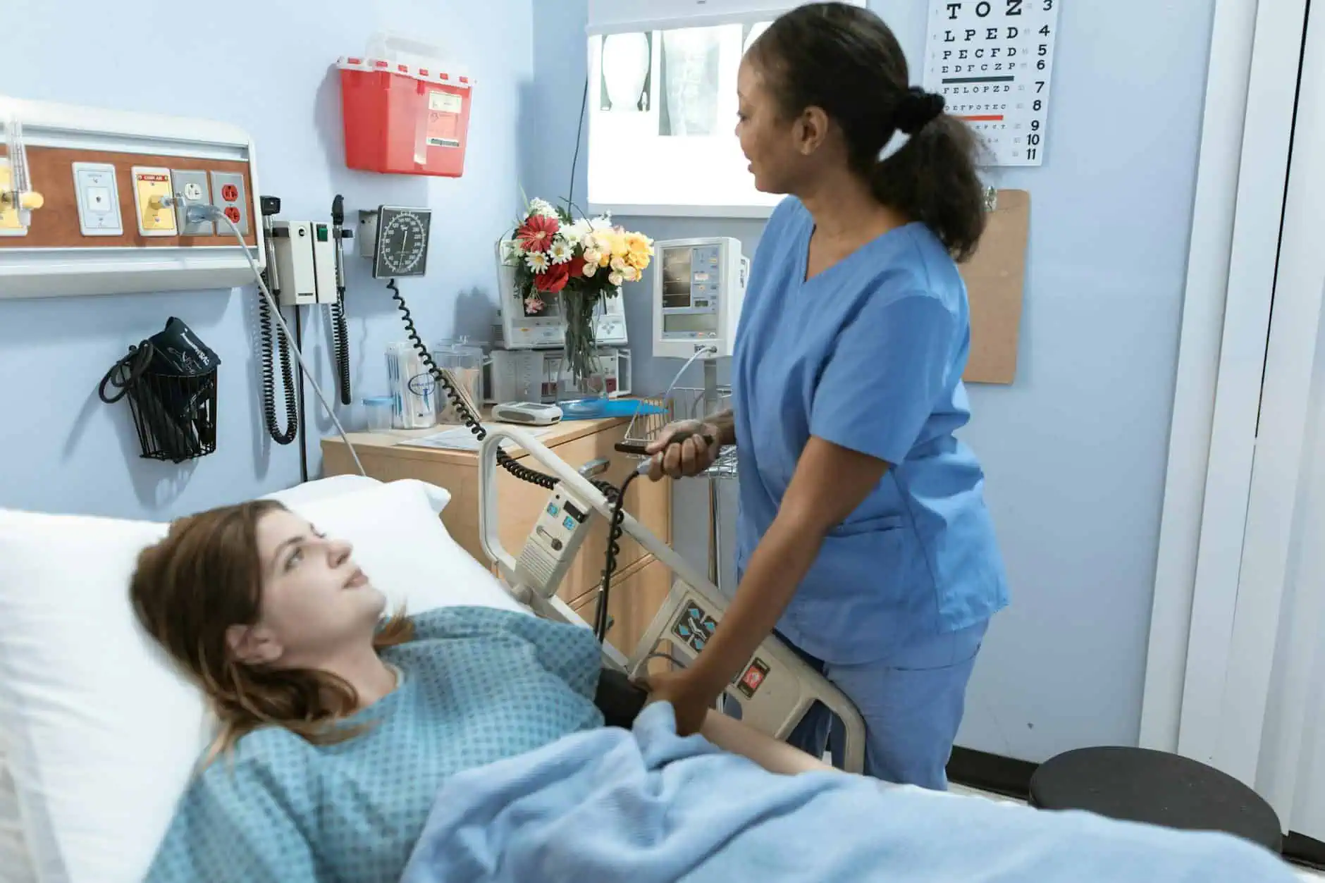 A doctor, dressed in blue scrubs, taking the blood pressure of a patient who is laying in a hospital bed.