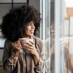 Stocksy_txp4044afb9FR2400_Medium_4211959 Stock photo of woman having a cup of coffee at home. She is smiling and looking through a window. She looks positive and happy.